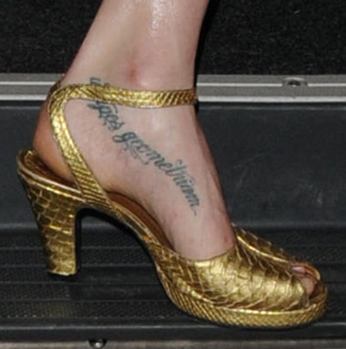 Lily Cole reveals her “Ut Apes Geometriam” foot tattoo in textured gold ankle-strap heels, with the sculpted silhouette and metallic finish highlighting the elegant Latin script along the inner arch