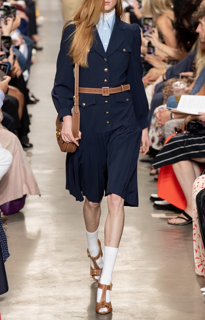 Michael Kors Collection’s pleated wool serge military dress delivers sharp tailoring with feminine movement, combining structured gold-button detailing and a belted waist with a softly pleated skirt for polished, modern elegance
