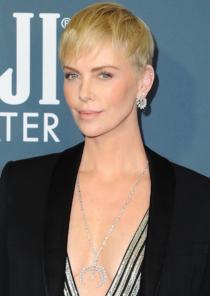 A close-up highlights Charlize’s sleek blonde pixie, understated peach-toned makeup, and crescent moon diamond necklace that adds quiet sparkle against the plunging neckline