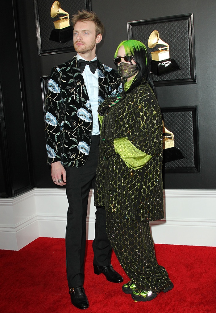 Finneas O’Connell and Billie Eilish celebrate their 2020 Grammy sweep in coordinated Gucci, with Finneas in a floral-embroidered tuxedo jacket and polished leather shoes, while Billie anchors her oversized monogram ensemble with chunky Flashtrek sneakers—blending classic tailoring with bold, street-driven maximalism
