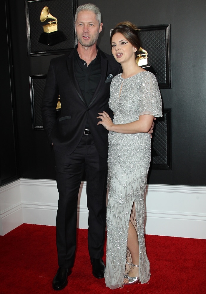 Lana Del Rey shows how her metallic Gucci knotted sandals echo the silver beadwork of her $595 Aidan Mattox gown alongside Sean “Sticks” Larkin at the 2020 Grammy Awards