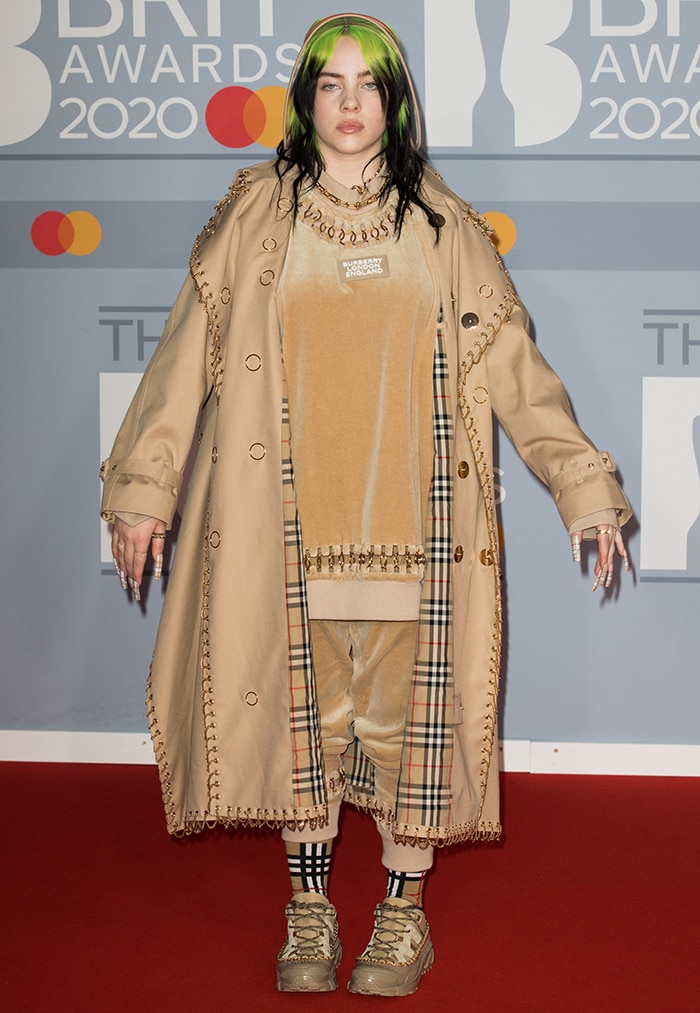 Billie Eilish arrives at the 2020 BRIT Awards in a custom Burberry look, layering an oversized chain-embellished trench over matching velour co-ords for a head-to-toe tonal statement