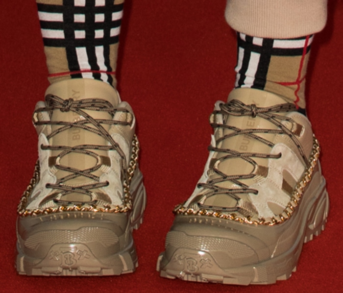 A closer look at her Burberry Arthur sneakers reveals exaggerated lug soles, hiking-style laces, and gold chain detailing that echo the hardware on her coat while grounding the oversized silhouette
