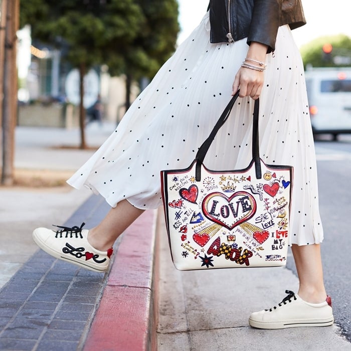 A playful Brighton tote covered in hand-drawn hearts and slogans adds a pop of personality to a breezy polka-dot skirt and casual sneakers