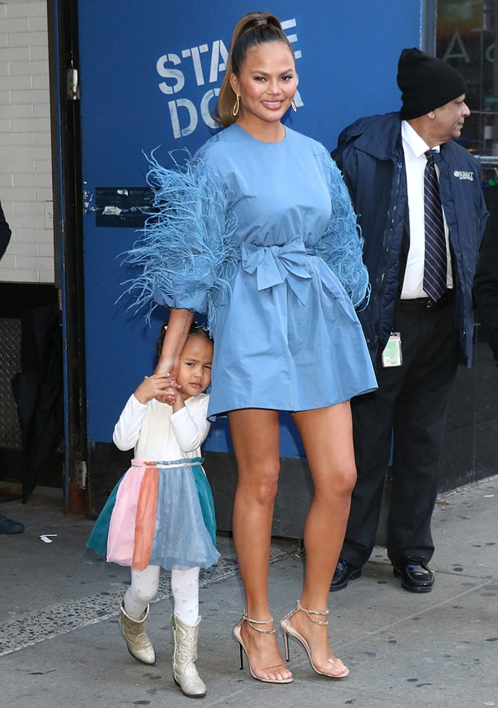 Chrissy Teigen arrives at the Strahan, Sara Keke studio in New York City on February 19, 2020, wearing a feather-trimmed Valentino mini dress styled with crystal-embellished Alevi Milano 'Valentina' sandals, while daughter Luna charms in a rainbow tulle dress and glittery cowboy boots