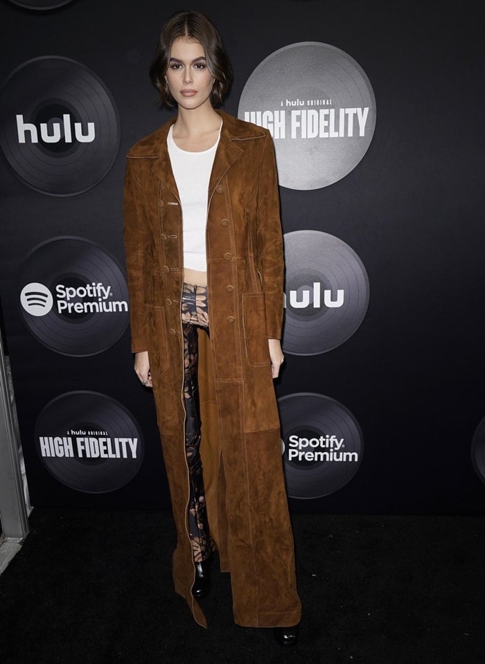 Kaia Gerber attends the premiere of Hulu's High Fidelity at Metrograph on February 13, 2020, in New York City, layering a sweeping brown suede Fendi trench coat over a white cropped top and Knwls' abstract-print Perse pants, finished with black leather ankle boots and Anita Ko diamond huggie earrings