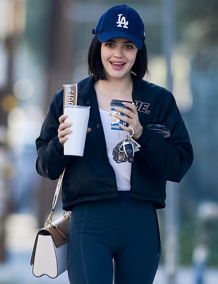 Carrying coffee and her Givenchy dog keyring, Lucy Hale softened the sharp lines of her monochrome athleisure look with a relaxed Dodgers cap and minimalist beauty styling