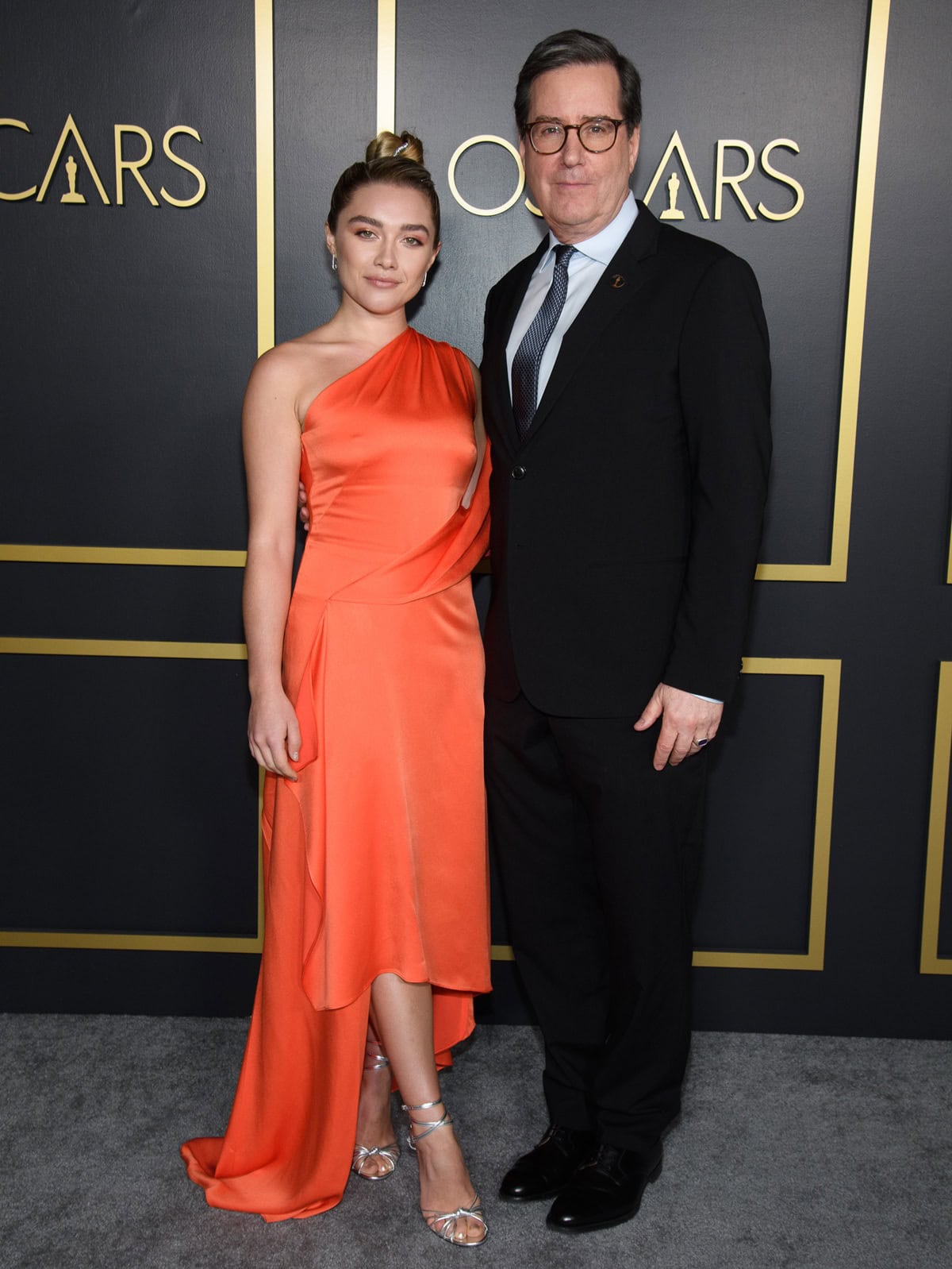 Oscar nominee Florence Pugh poses with Academy President David Rubin at the 92nd Oscars Nominees Luncheon, pairing her vibrant Monse satin dress with metallic silver Jimmy Choo Lovella sandals that add cool contrast to the citrus-orange ensemble