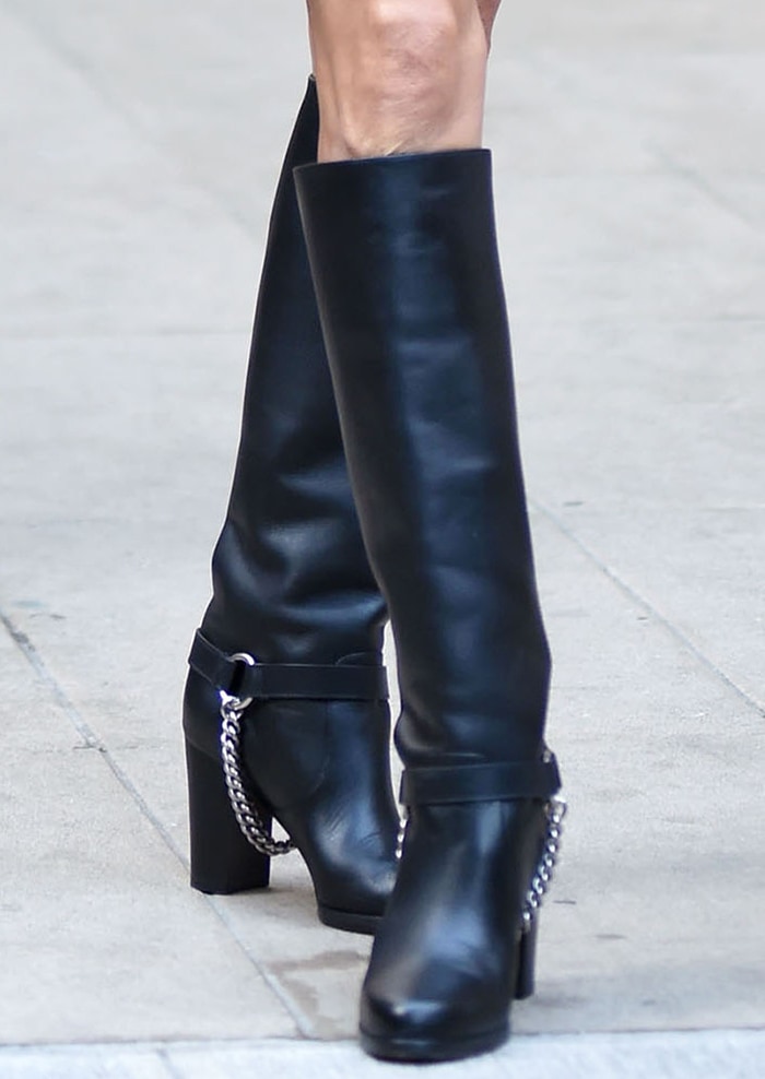 Celine Dion grounds her Michael Kors cape dress with sleek black leather riding boots featuring chunky block heels and silver chain accents for a polished equestrian finish