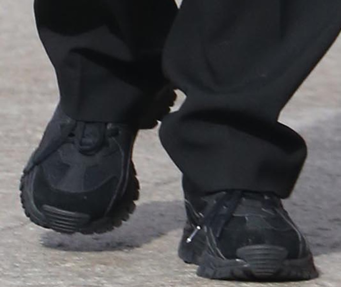 A close-up reveals the all-black athletic sneakers’ rugged tread, layered construction, and technical detailing beneath Emily’s floor-skimming trousers