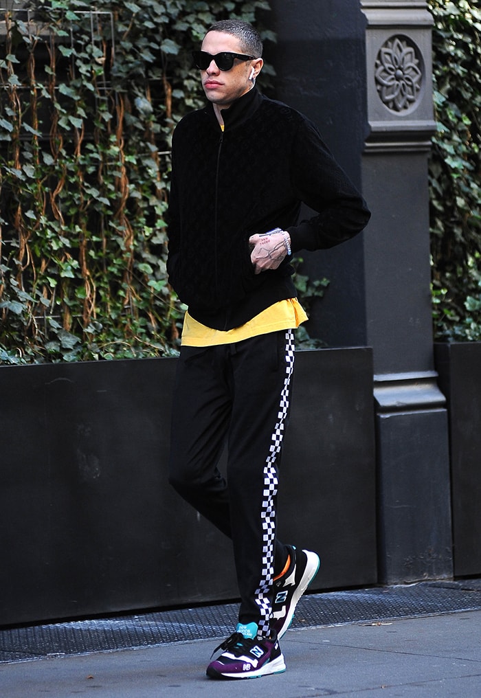 Pete Davidson wears colorful New Balance sneakers with checkerboard track pants and a black jacket while leaving Kaia Gerber’s SoHo apartment in October 2019
