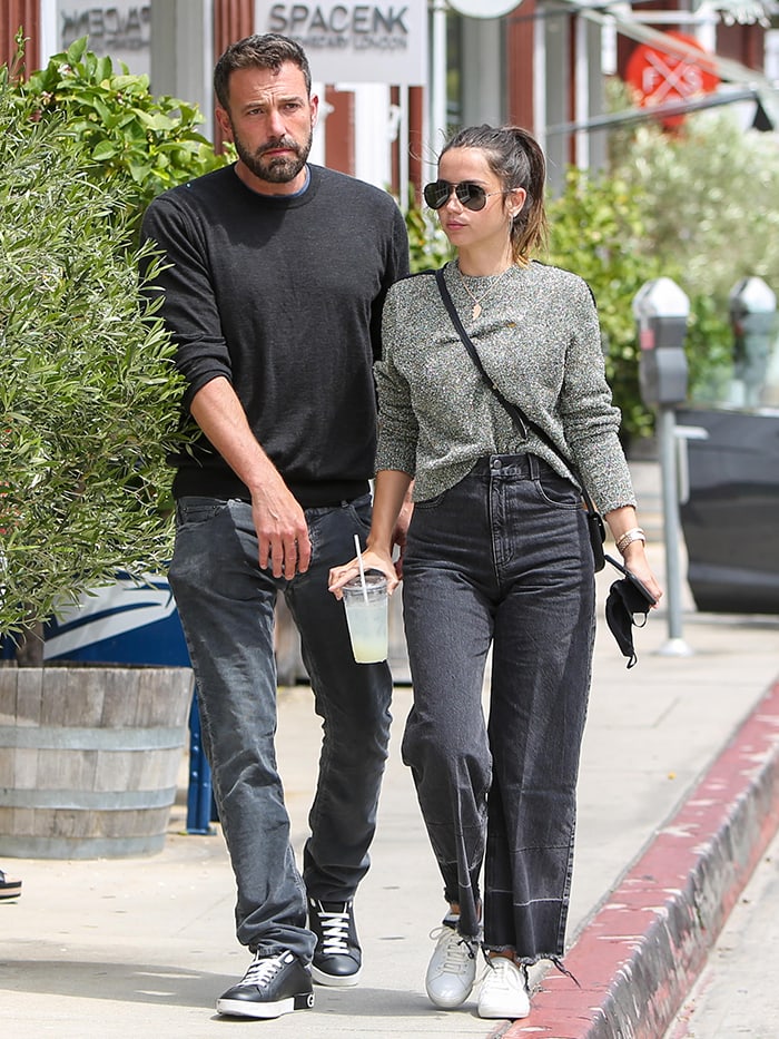 Ana de Armas embraces understated off-duty style in a Bella Freud metallic-knit sweater, Rachel Comey washed-black denim pants, and white Saint Laurent “Andy” sneakers, while Ben Affleck keeps things simple in a black crewneck sweater, faded jeans, and black low-top sneakers