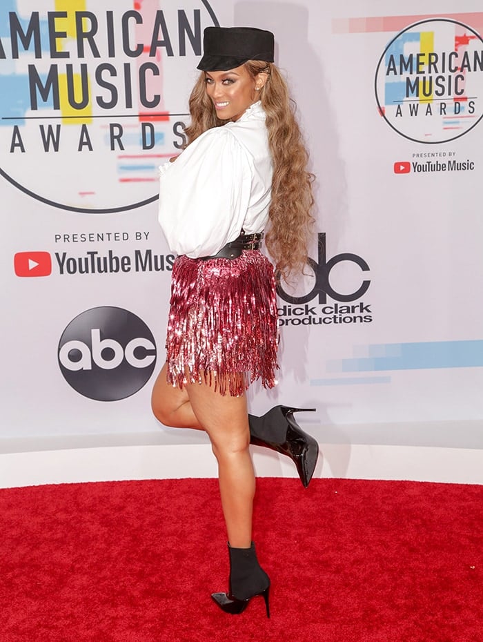 Tyra Banks towers in a playful pose at the 2018 American Music Awards, wearing a confetti-fringed Gucci Fall 2018 ensemble with black sock boots—though her accessories and styling choices somewhat undercut the look’s high-fashion potential