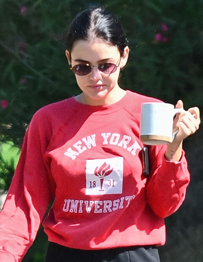 Lucy Hale keeps her morning look relaxed and collegiate, pairing a vintage red NYU sweatshirt with slim black shorts, round sunglasses, and a coffee mug in hand
