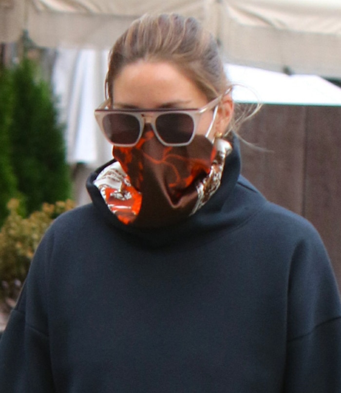 Olivia Palermo layers oversized sunglasses over a richly printed red-and-brown scarf, adding color and pattern to her otherwise all-black athleisure look