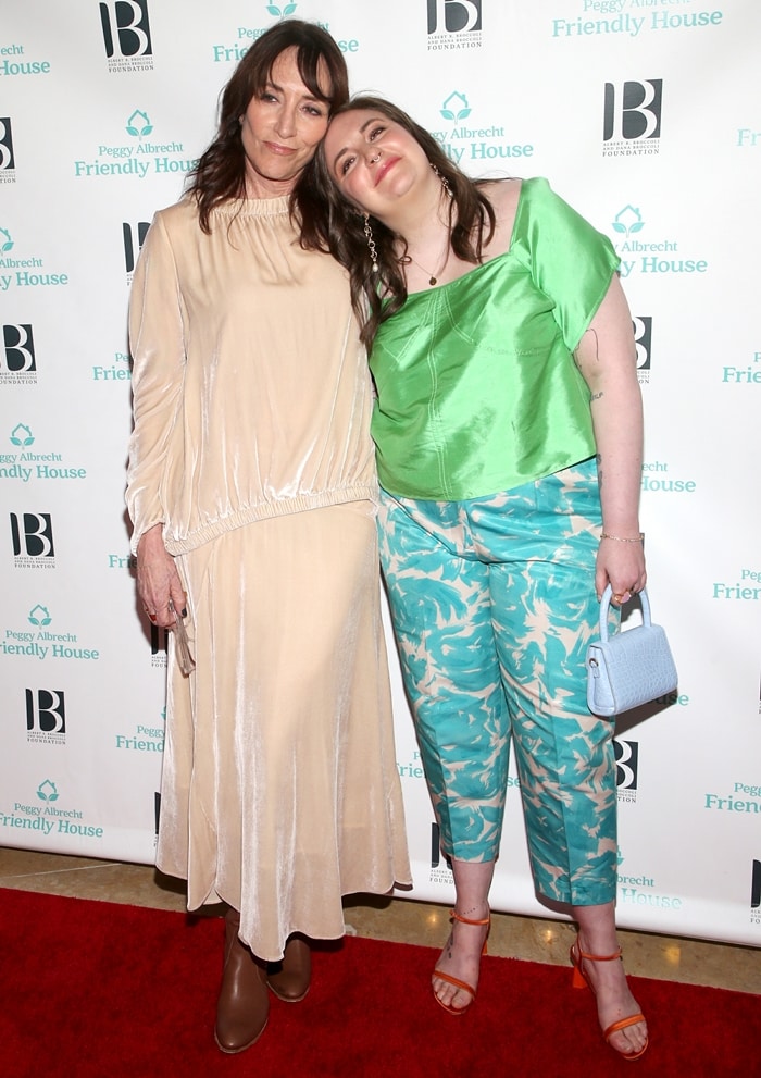 Lena Dunham pairs vibrant orange ankle-strap heels with tropical print pants and a satin green top, standing beside Katey Sagal in a neutral-toned velvet midi dress and tan boots at the Peggy Albrecht Friendly House Awards