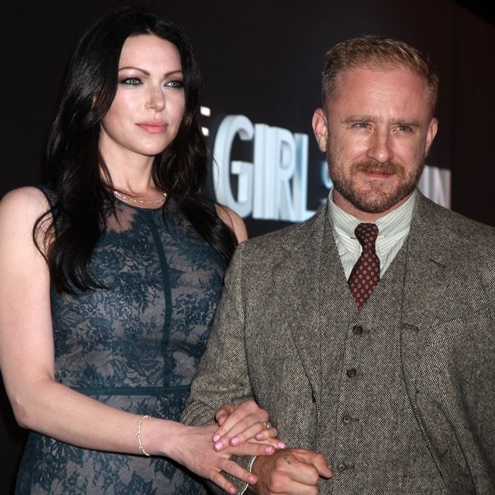 Laura Prepon towers over her husband Ben Foster as they pose together—she exudes elegance in a sheer navy lace dress, while Foster keeps it classic in a textured three-piece brown tweed suit paired with a maroon patterned tie and striped shirt