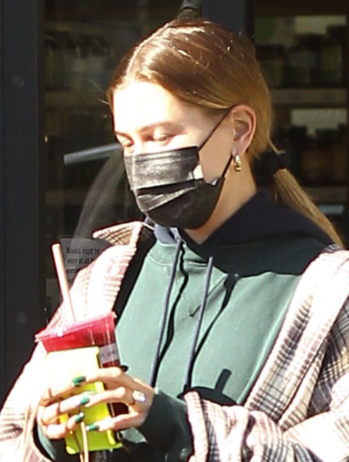 Hailey Bieber carries a smoothie while sporting gold hoop earrings, green nail polish, and a relaxed ponytail that complements her laid-back athleisure look