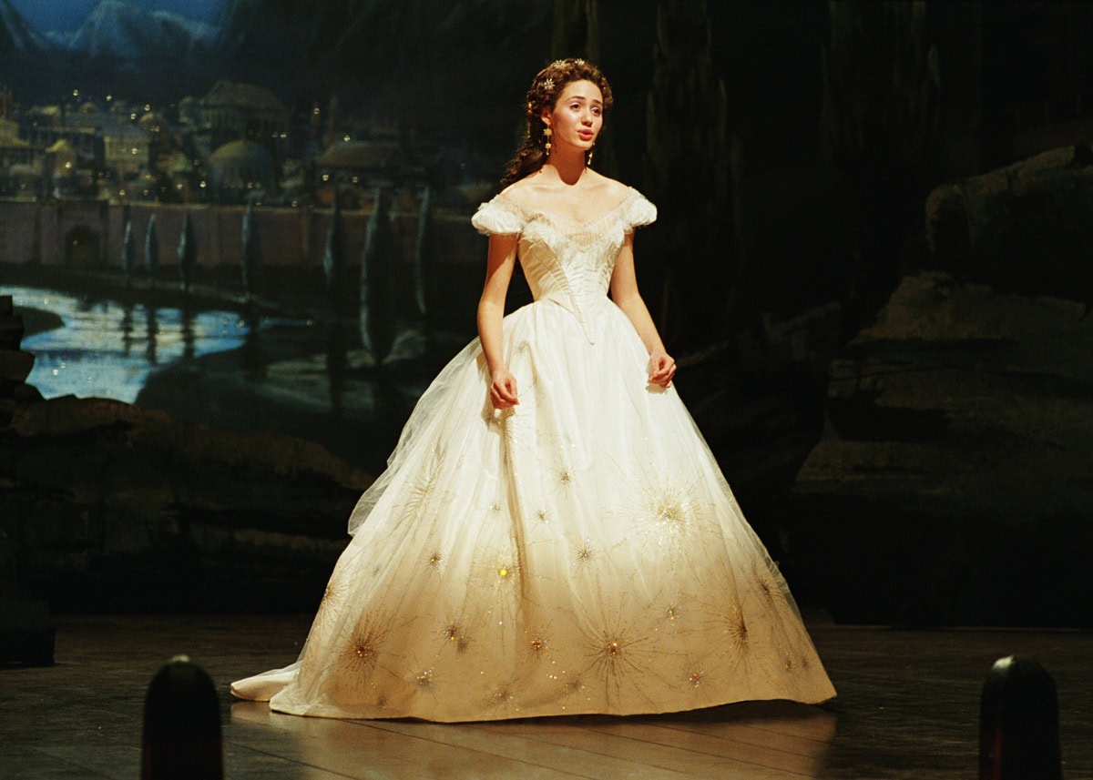 Emmy Rossum wears the iconic off-the-shoulder white ballgown as Christine Daaé in the “Think of Me” scene from The Phantom of the Opera (2004), with intricate starburst embroidery by costume designer Alexandra Byrne