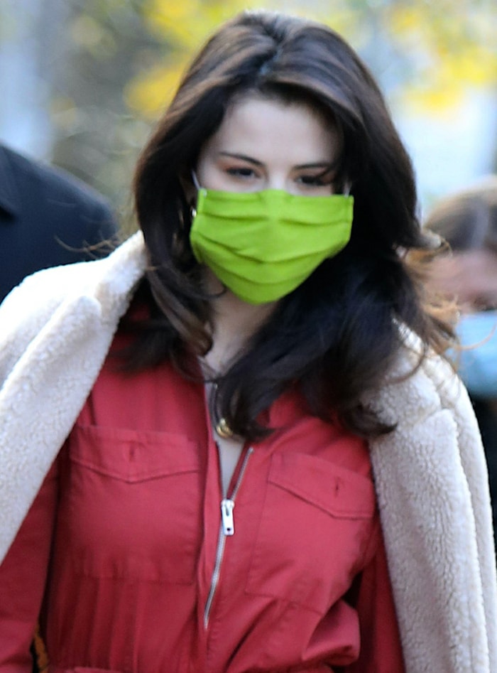 Selena Gomez showcases soft, voluminous waves and a lime-green face mask, adding a playful pop of color to her red jumpsuit on the “Only Murders in the Building” set