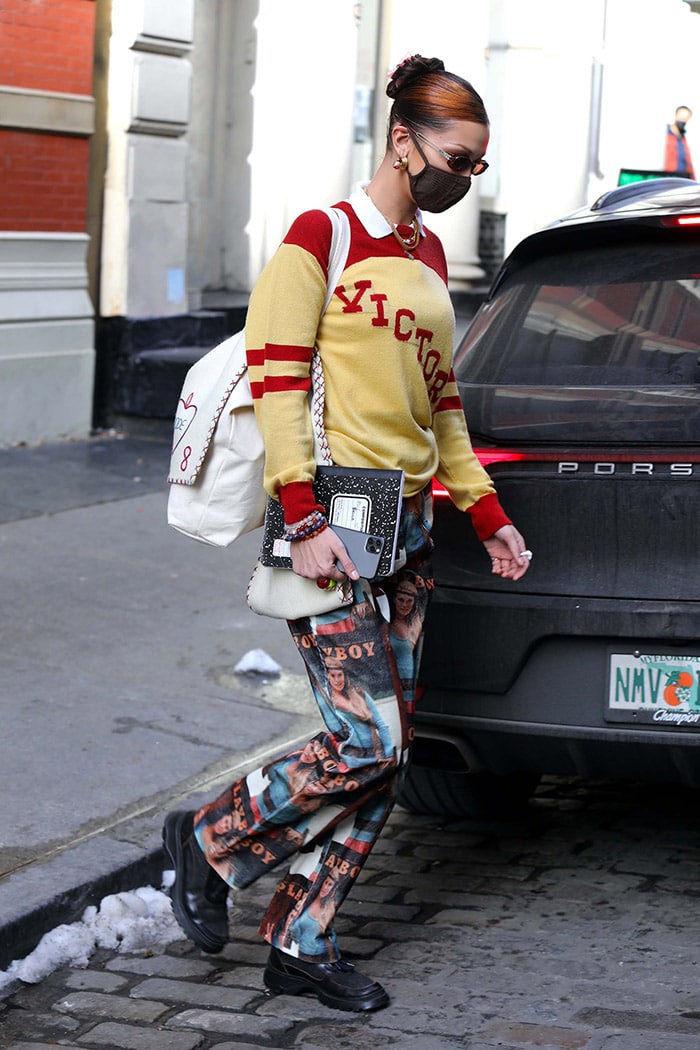 Bella Hadid elevates her collegiate-inspired look with an Emi Jay Papillon clip in Sugar Plum, a Bode Victoria sweater, a Skims seamless mask, layered Monica Vinader Doina and Bvlgari ruby-diamond necklaces, and Soulland’s retro Playboy Fawna pants