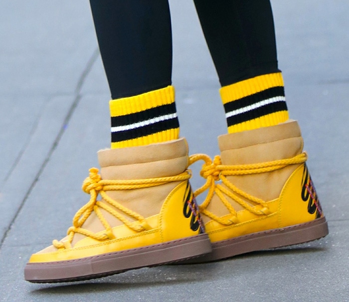 A close-up of Olivia Palermo's bright yellow Inuikii shearling sneaker boots showcases their suede uppers, rope laces, and gum soles, paired with striped athletic socks