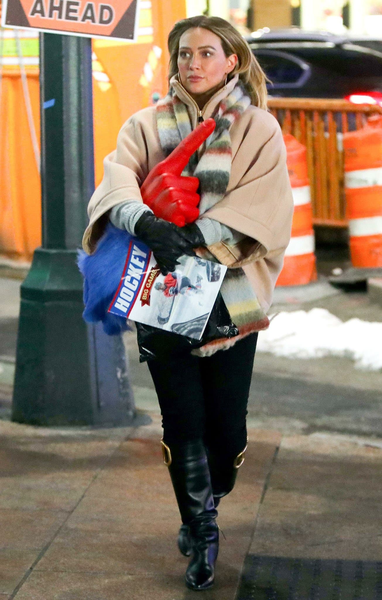 Hilary Duff layers a blush coat and striped scarf over black trousers, finishing the winter-ready Younger ensemble with Valentino Garavani Supervee knee-high boots featuring oversized gold-tone V-logo hardware