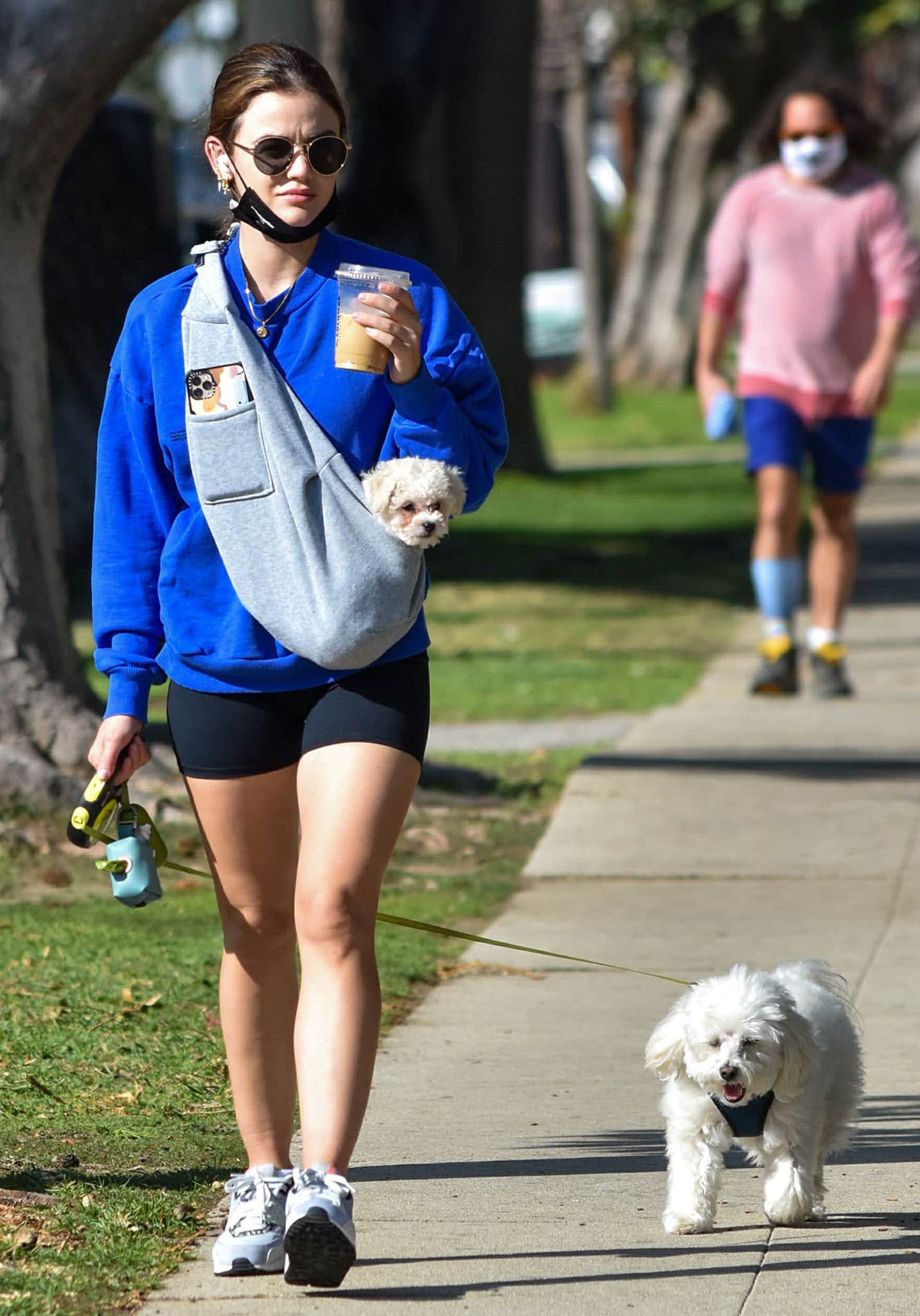 Lucy Hale walks Elvis while carrying newly adopted rescue dog Ethel in a sling, pairing sneakers with a cobalt-blue Pangaia sweatshirt and black cycling shorts