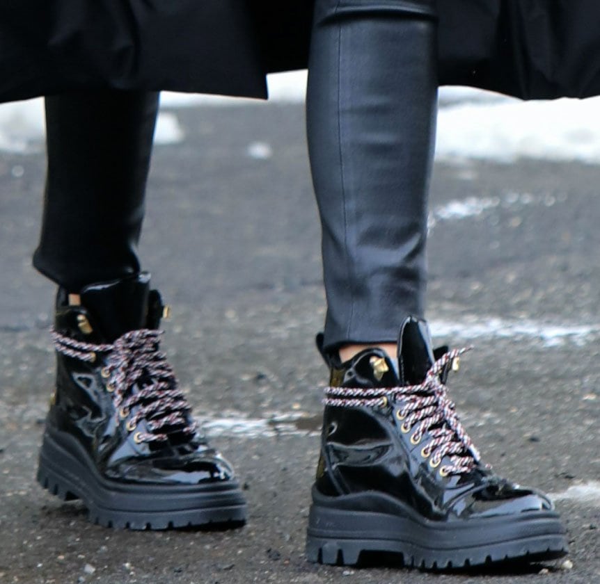 A close-up of Olivia Palermo's glossy Fratelli Rossetti patent leather ankle boots showcases their chunky lug soles, contrasting hiking-style laces, gold-tone eyelets, and rugged silhouette paired with sleek leather leggings