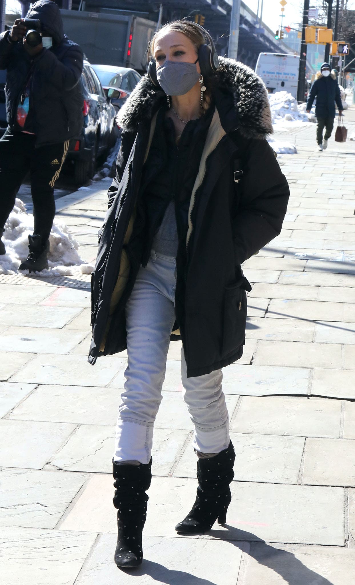 Sarah Jessica Parker steps out in New York City on February 10, 2021, bundled in a Woolrich Arctic Down parka with white jeans, finishing the winter-ready look with studded SJP by Sarah Jessica Parker “Busker” boots, a Proper Cloth grey face mask with a Pretty Connected chain, Beats Studio3 headphones, and her Samsonite x SJP “Carried Away” convertible bag