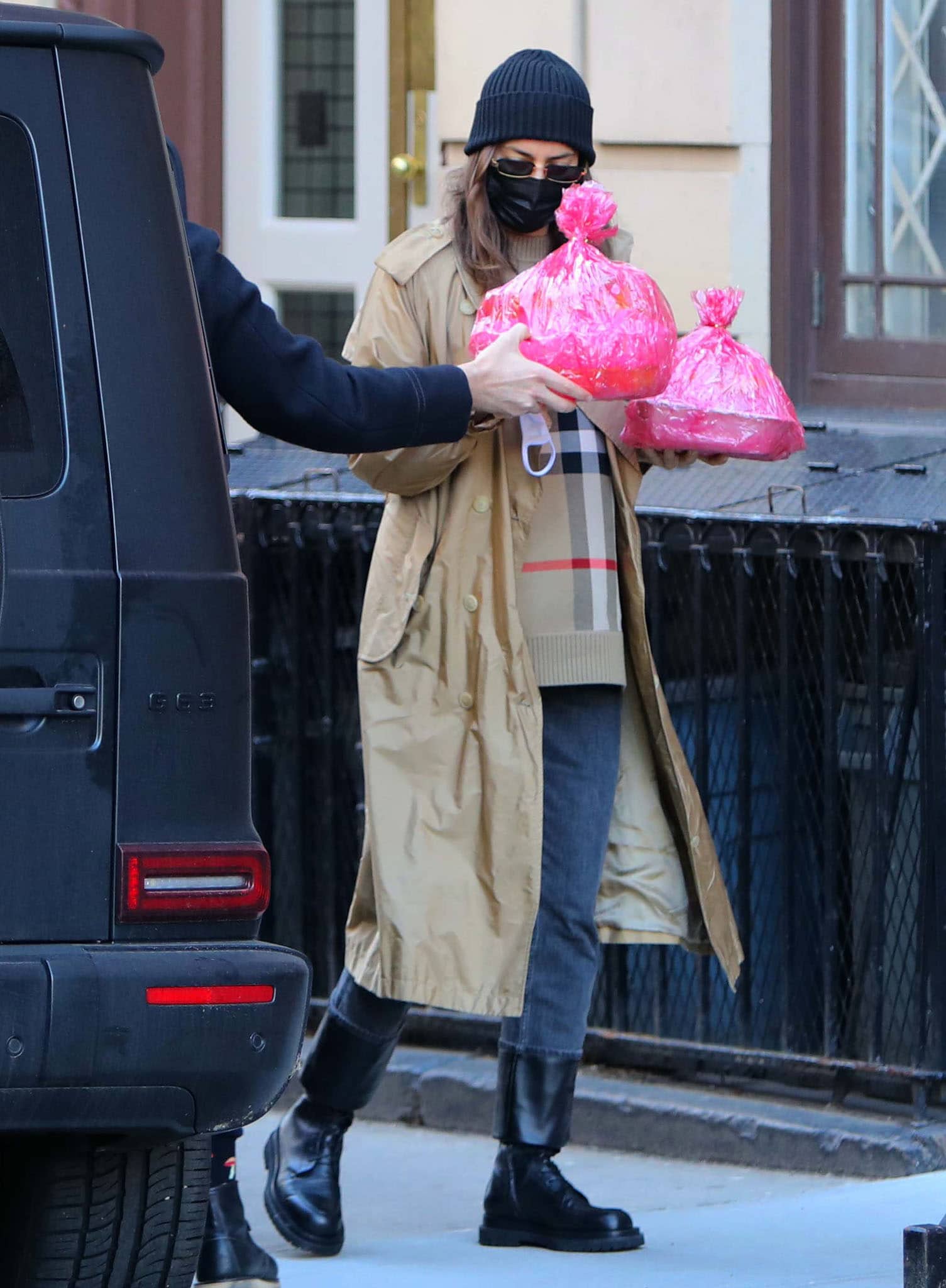 Irina Shayk carries pink-wrapped birthday treats in a Burberry trench and check sweater, grounding the layered look with black Magda Butrym combat boots