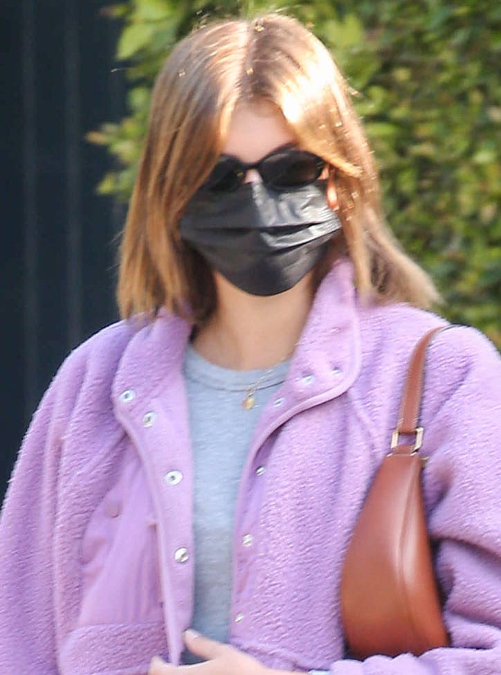 Kaia Gerber pairs a plush lavender fleece jacket with a gray cropped top, finishing the look with slim black sunglasses, a black face mask, and a tan shoulder bag tucked under her arm