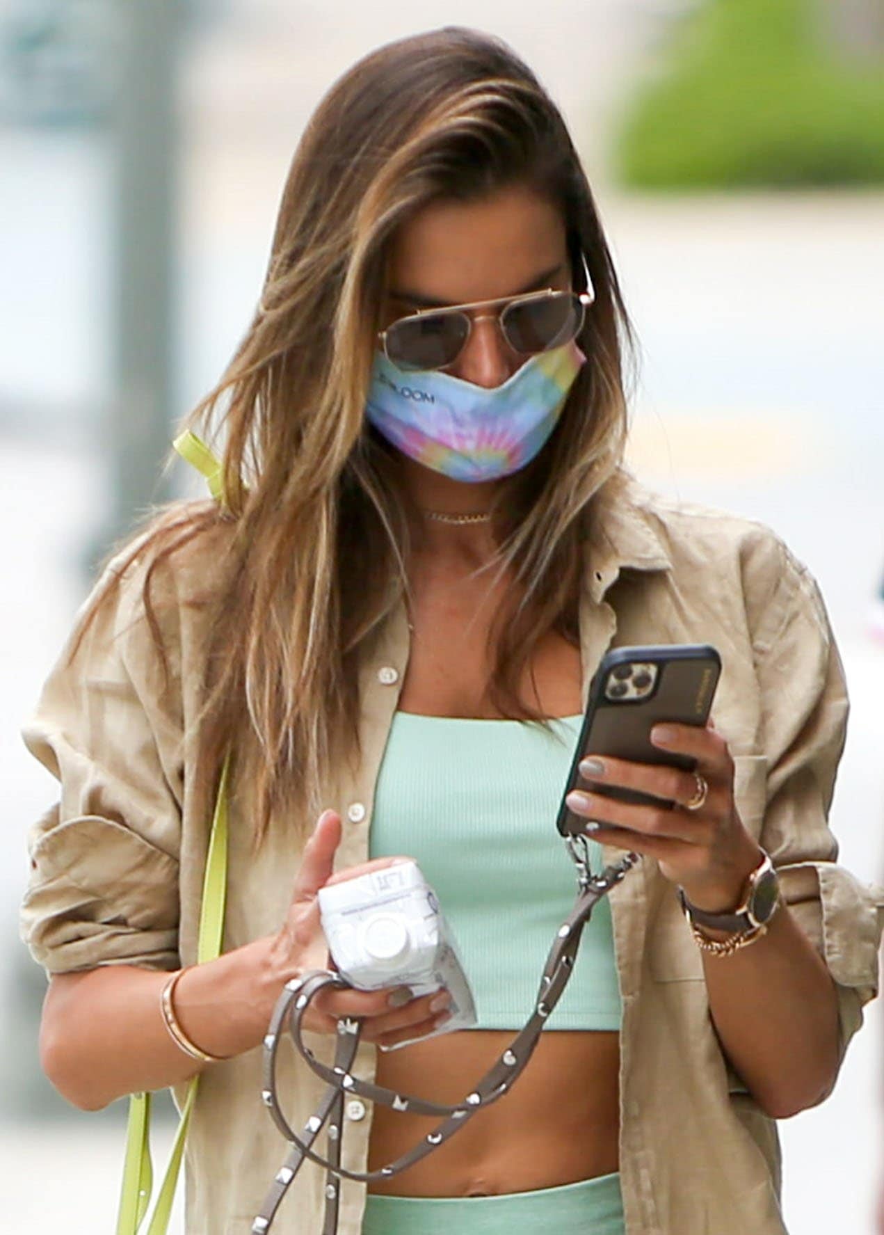 Alessandra Ambrosio layers a beige linen shirt over a honeydew-green Alo Yoga set, accessorizing with aviator sunglasses, gold jewelry, and a tie-dye face mask