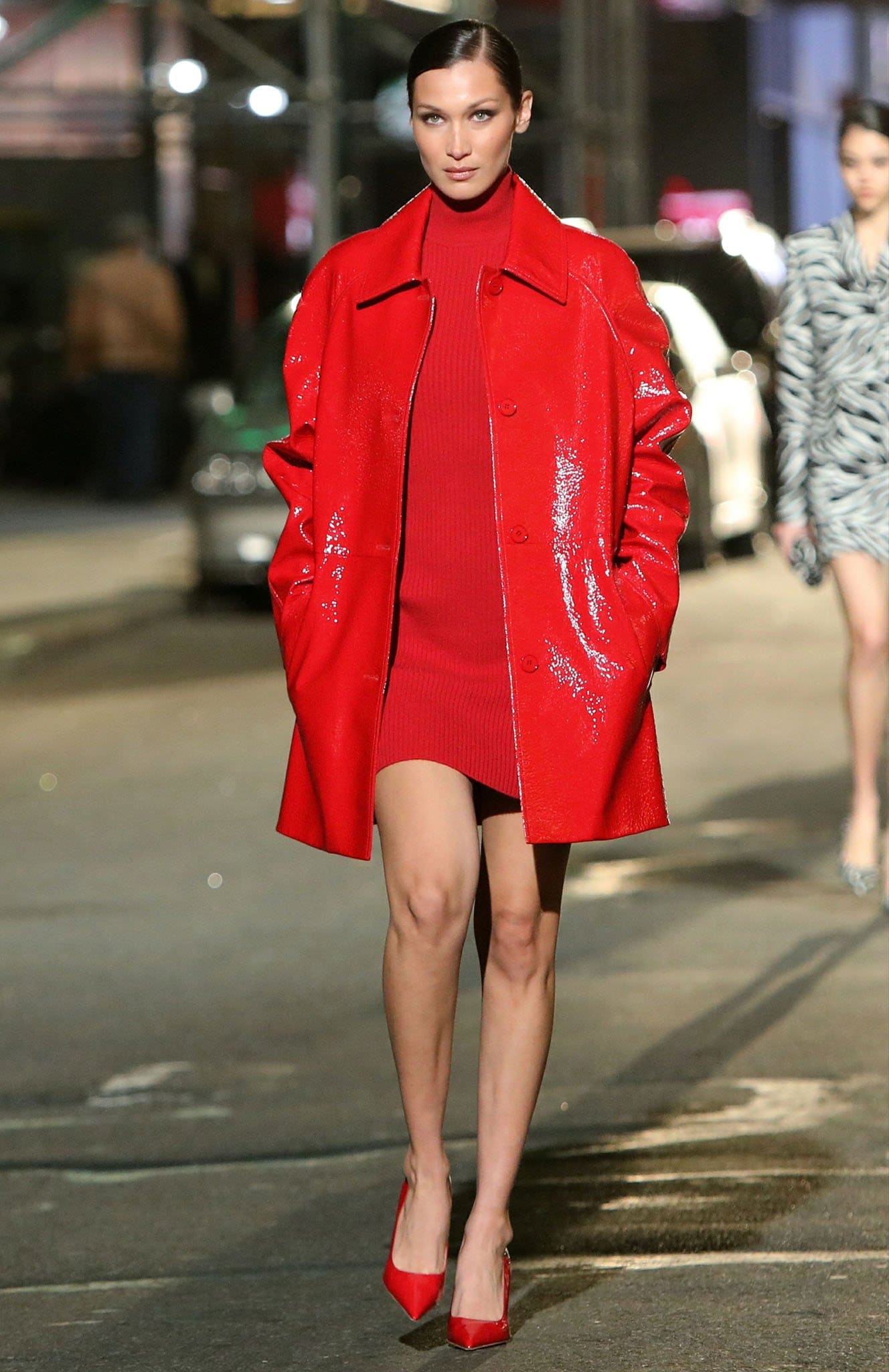 Bella Hadid wears a monochromatic scarlet look featuring a ribbed knit mini dress, glossy patent leather trench coat, and matching pointed-toe pumps