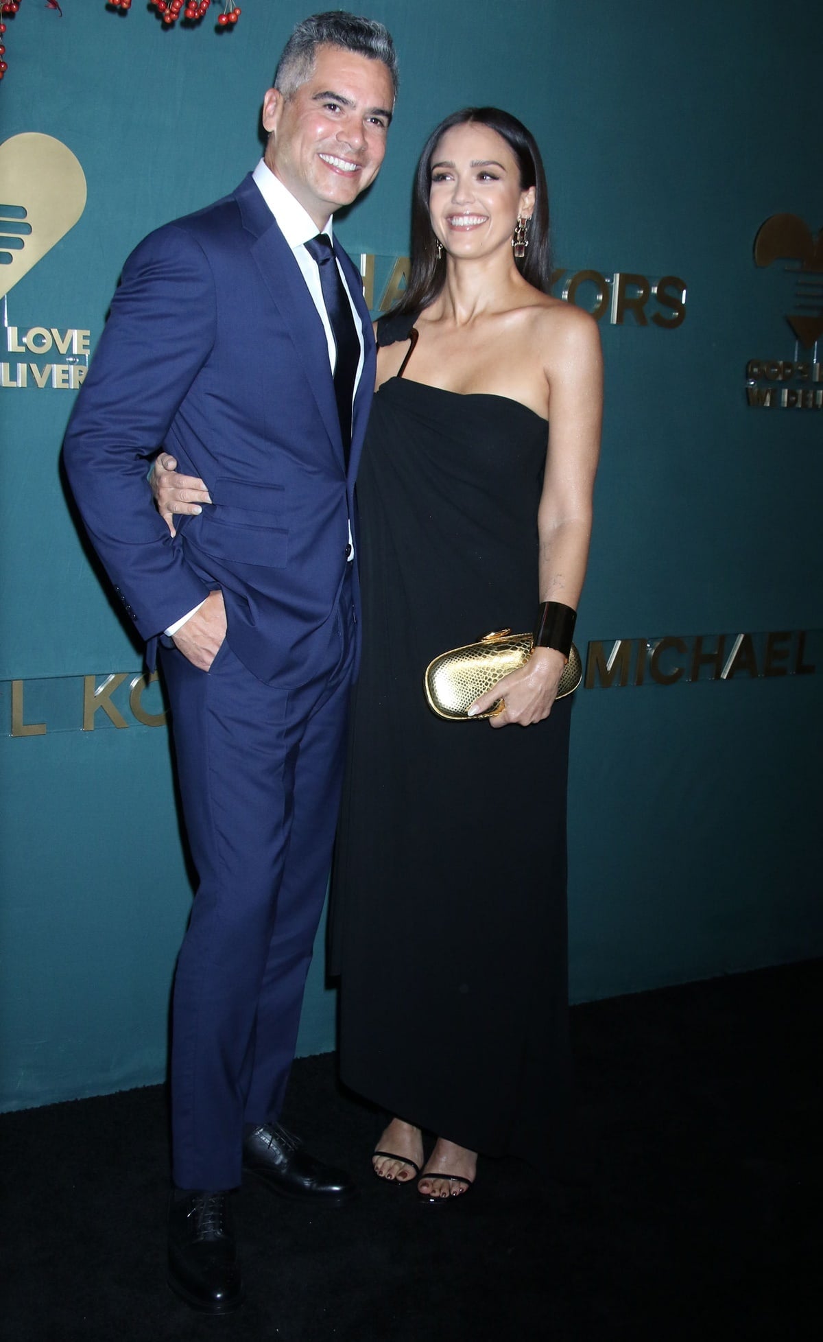 Jessica Alba glowed on the black carpet in a sleek one-shoulder Michael Kors Collection gown, pairing the look with strappy Stuart Weitzman sandals, a gold clutch, and sculptural jewelry while posing alongside her husband, Cash Warren