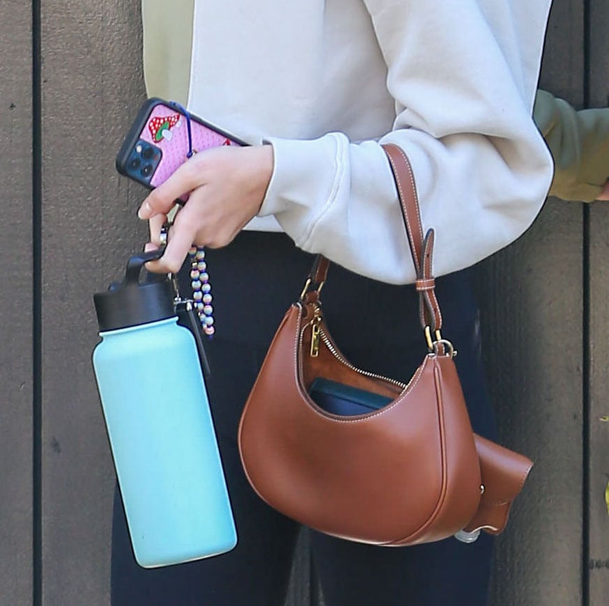 Kaia Gerber accessorizes her off-duty look with a curved Celine shoulder bag, pairing it with a pastel Simple Modern Summit water bottle and an iPhone tucked in a playful Wildflower Shrooms case for a subtle mix of polish and personality