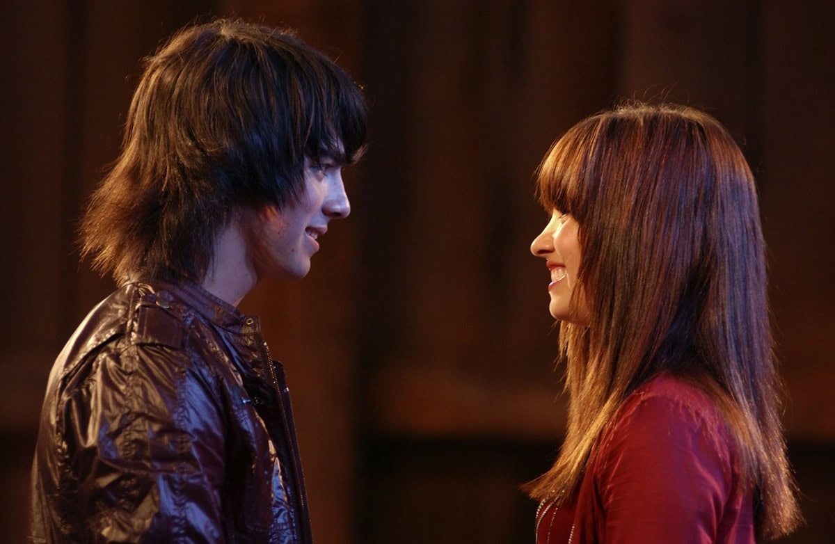 Joe Jonas as Shane Gray and Demi Lovato as Mitchie Torres share a heartfelt moment in Camp Rock, both dressed in coordinating red-toned jackets that reflect their budding on-screen connection