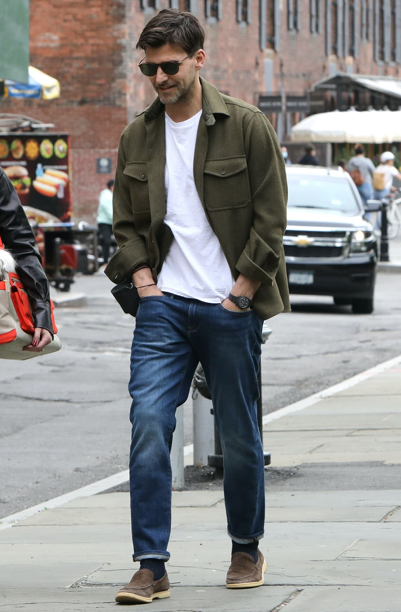 Johannes Huebl keeps his Dumbo look casual in cuffed blue jeans, an army-green jacket, brown suede loafers, and dark sunglasses