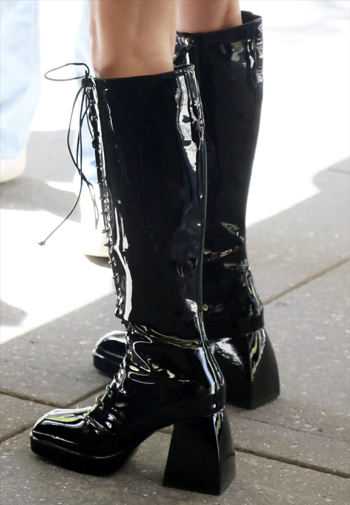 Olivia Rodrigo steps out in glossy black knee-high PVC boots featuring a squared toe, lace-up front with side zip closure, and a chunky flared block heel that adds height and a bold, retro-inspired edge to her look