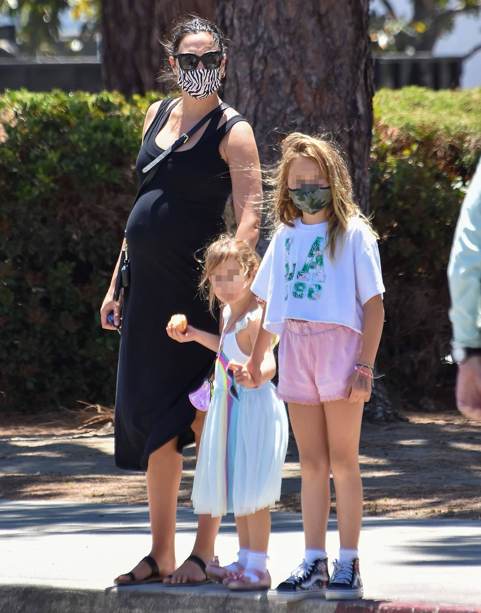 Pregnant Gal Gadot pairs a sleeveless black jersey midi dress with minimalist toe-ring sandals, a Bandolier crossbody phone case, gold hoop earrings, and a zebra-print face mask