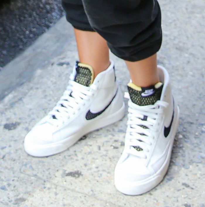 Olivia Culpo completes her sporty NYC look with classic Nike Blazer Mid sneakers, featuring crisp white leather uppers, bold black Swooshes, and mesh tongues accented with neon green and yellow detailing
