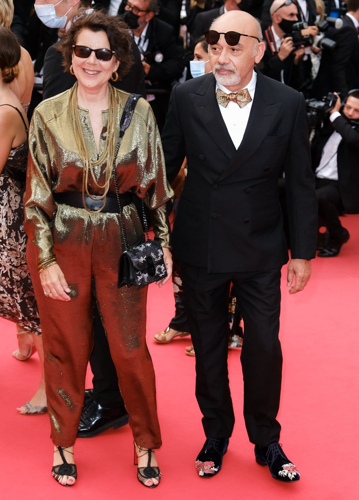 Christian Louboutin (R) steps onto the Cannes red carpet in embroidered velvet shoes and a sharp tuxedo, joined by a guest in a metallic gold jumpsuit, at the premiere of The French Dispatch during the 74th annual Cannes Film Festival on July 12, 2021