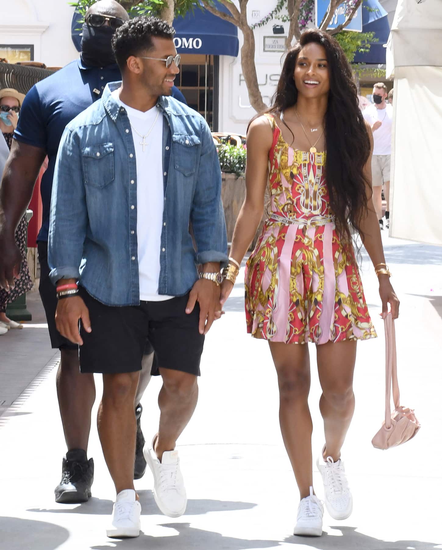 Ciara smiles as she walks beside Russell, her vibrant Moschino mini dress contrasting with clean white Nike sneakers for a polished yet casual finish