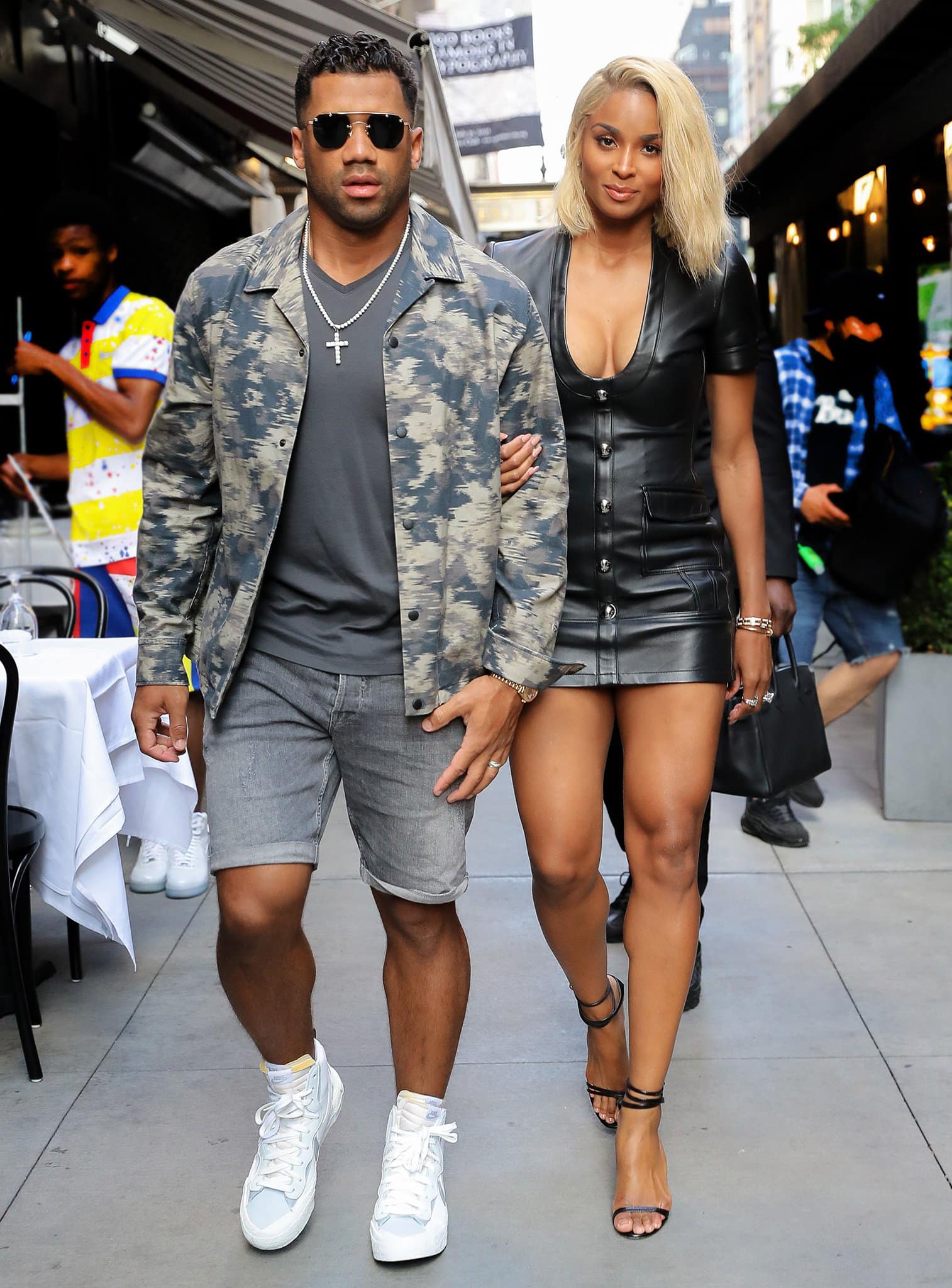 Ciara highlights her toned legs in a plunging David Koma black leather mini dress styled with sleek black ankle-strap stiletto sandals, while Russell Wilson keeps it cool beside her in a camo jacket, charcoal tee, denim shorts, and crisp white Nike sneakers