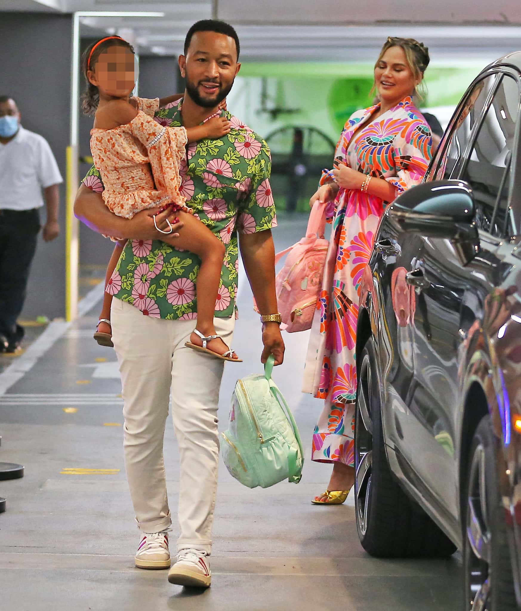 John Legend pairs his green floral Hawaiian shirt with crisp white trousers and classic low-top sneakers while carrying Luna in delicate white ankle-strap sandals