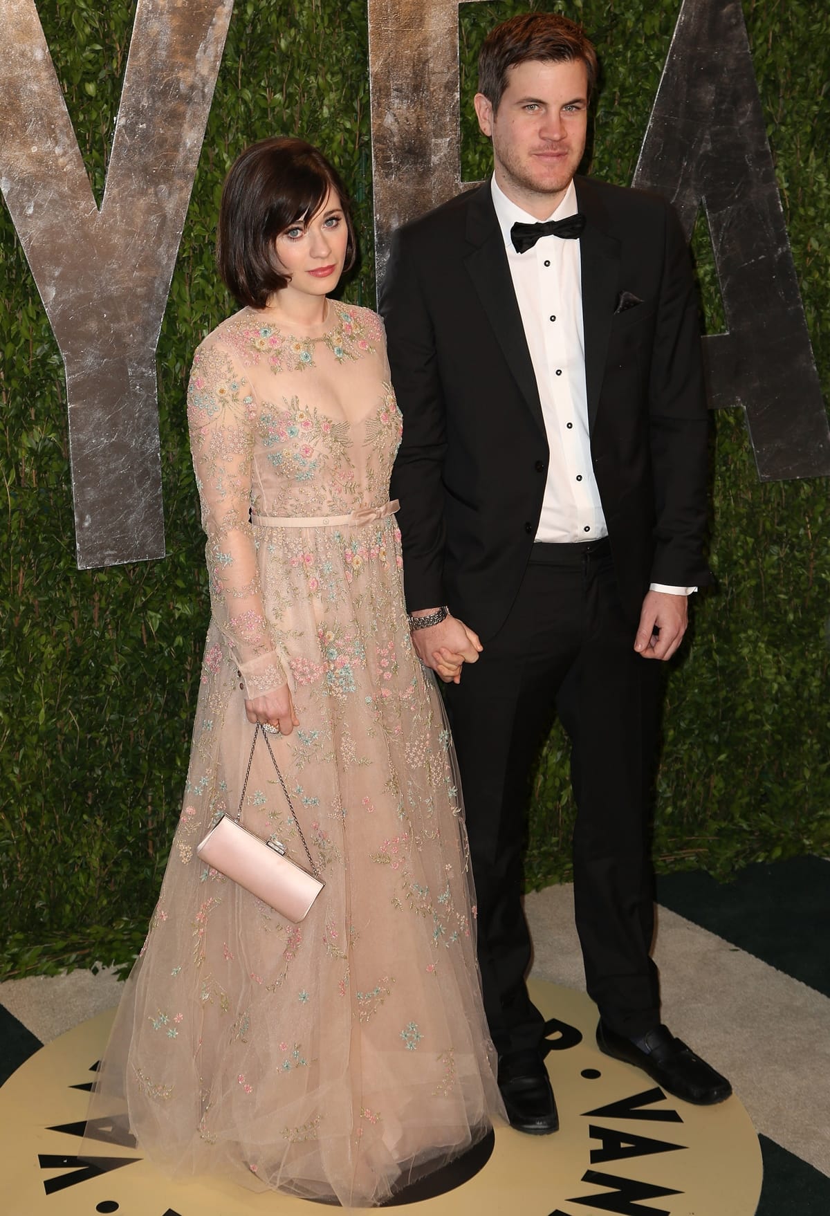Zooey Deschanel looks ethereal in a blush pink floral-embroidered gown with sheer sleeves as she attends the Vanity Fair Oscar Party hand-in-hand with screenwriter Jamie Linden