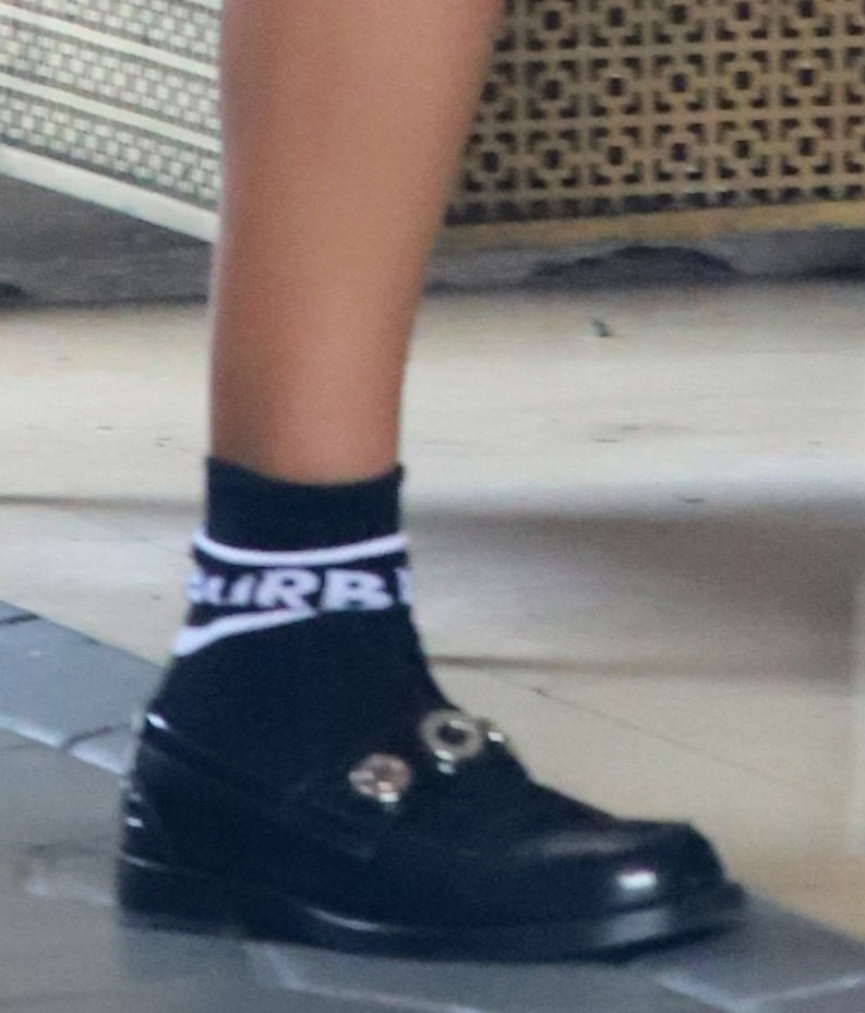 A close-up reveals glossy black loafers accented with gold hardware, grounded by Burberry logo socks that add a sporty edge to the tailored silhouette