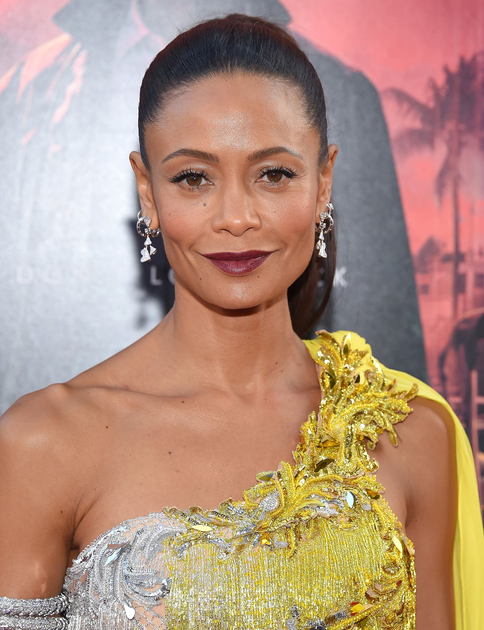 Thandiwe Newton stuns at the Los Angeles premiere of Reminiscence, wearing a one-shoulder silver and yellow gown adorned with intricate embroidery and fringe detailing, complemented by a sleek ponytail and deep plum lipstick