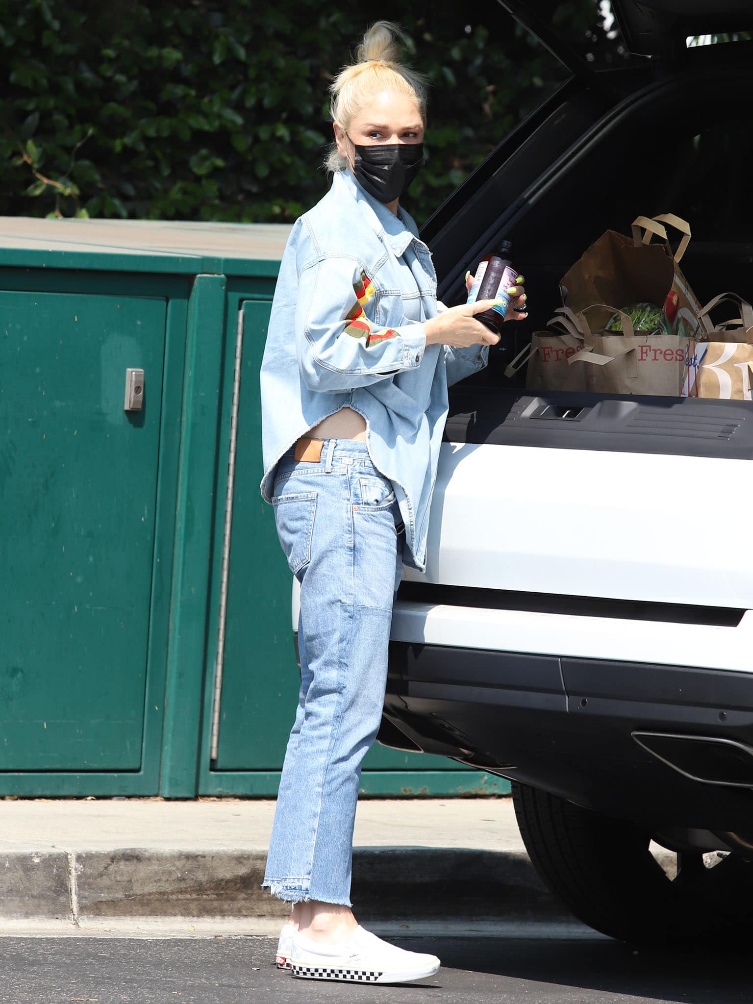 Gwen Stefani loads groceries into her SUV while showcasing a relaxed denim-on-denim look paired with checkerboard Vans Slip-On SF sneakers and a casual top-knot hairstyle