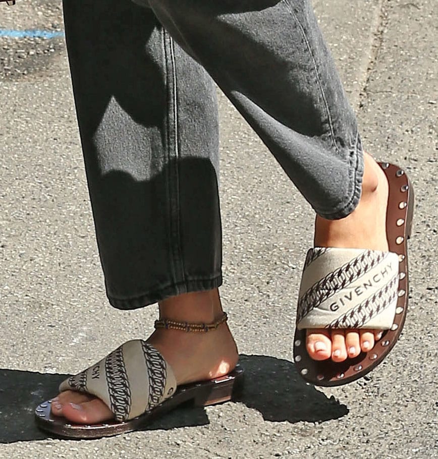 Sofia Richie grounds her minimalist look with logo-jacquard Givenchy slide sandals, featuring a padded monogram strap and a subtly stacked sole trimmed with metal studs—an elevated take on the classic flat that adds texture and quiet designer edge to her off-duty ensemble